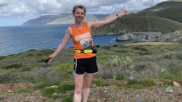 Kate McCurdy, wearing a Fred’s Team tank top, stretches her arms out wide, with her back turned to a lake surrounded by mountains. 