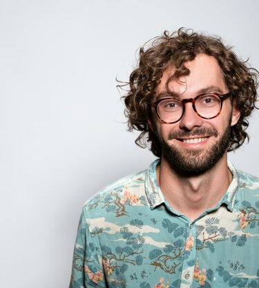 Smiling man with curly hair and glasses, wearing a patterned shirt.