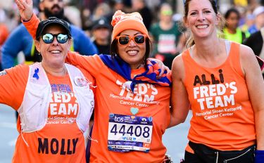 Three Fred's Team charity runners wearing orange Fred’s Team uniforms stand shoulder to shoulder during the TCS New York City Marathon.