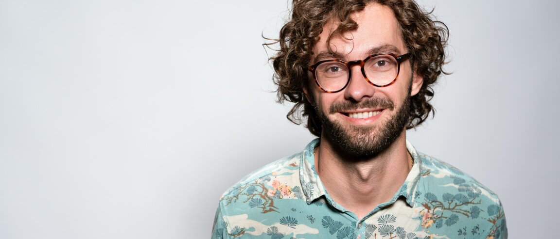 Smiling man with curly hair and glasses, wearing a patterned shirt.