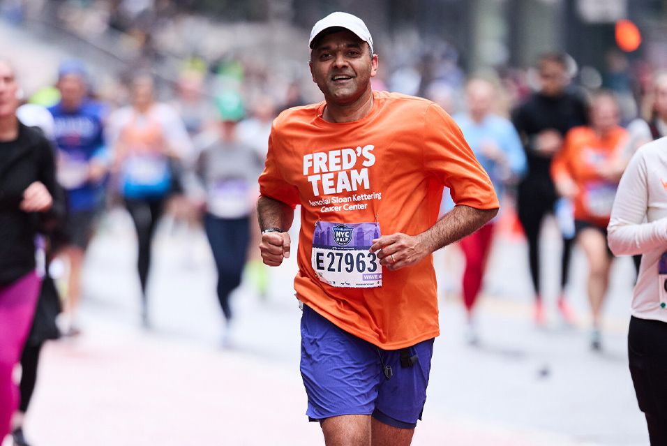 A runner in an orange Fred’s Team shirt jogs on a New York City street amid a crowd of marathon participants. 
