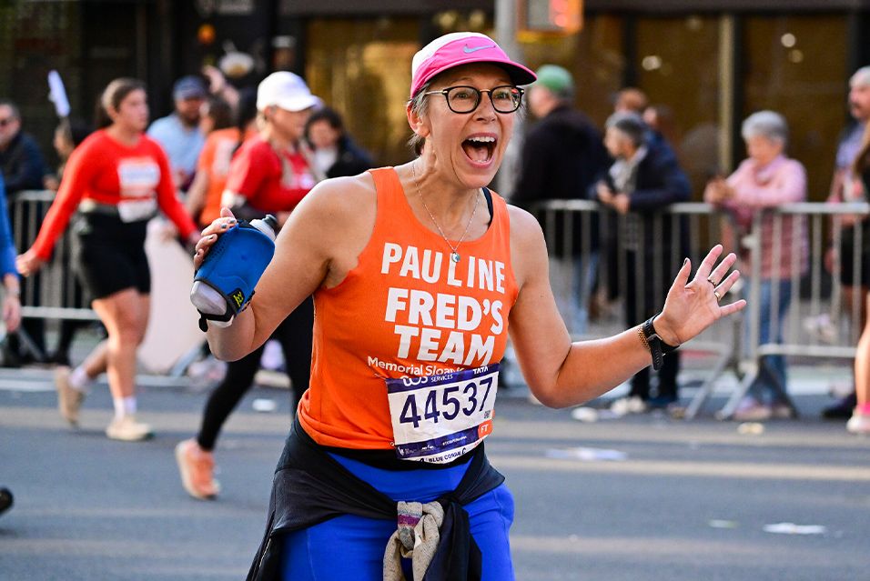 A runner wearing a Fred's Team tank top and a pink cap moves forward with arms raised, holding a water bottle.  