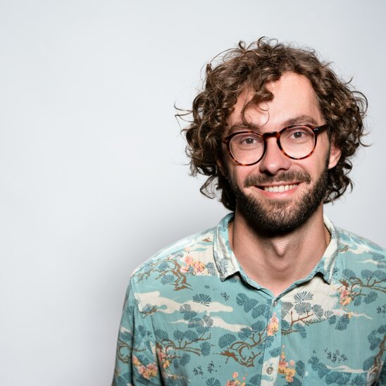 Smiling man with curly hair and glasses, wearing a patterned shirt.